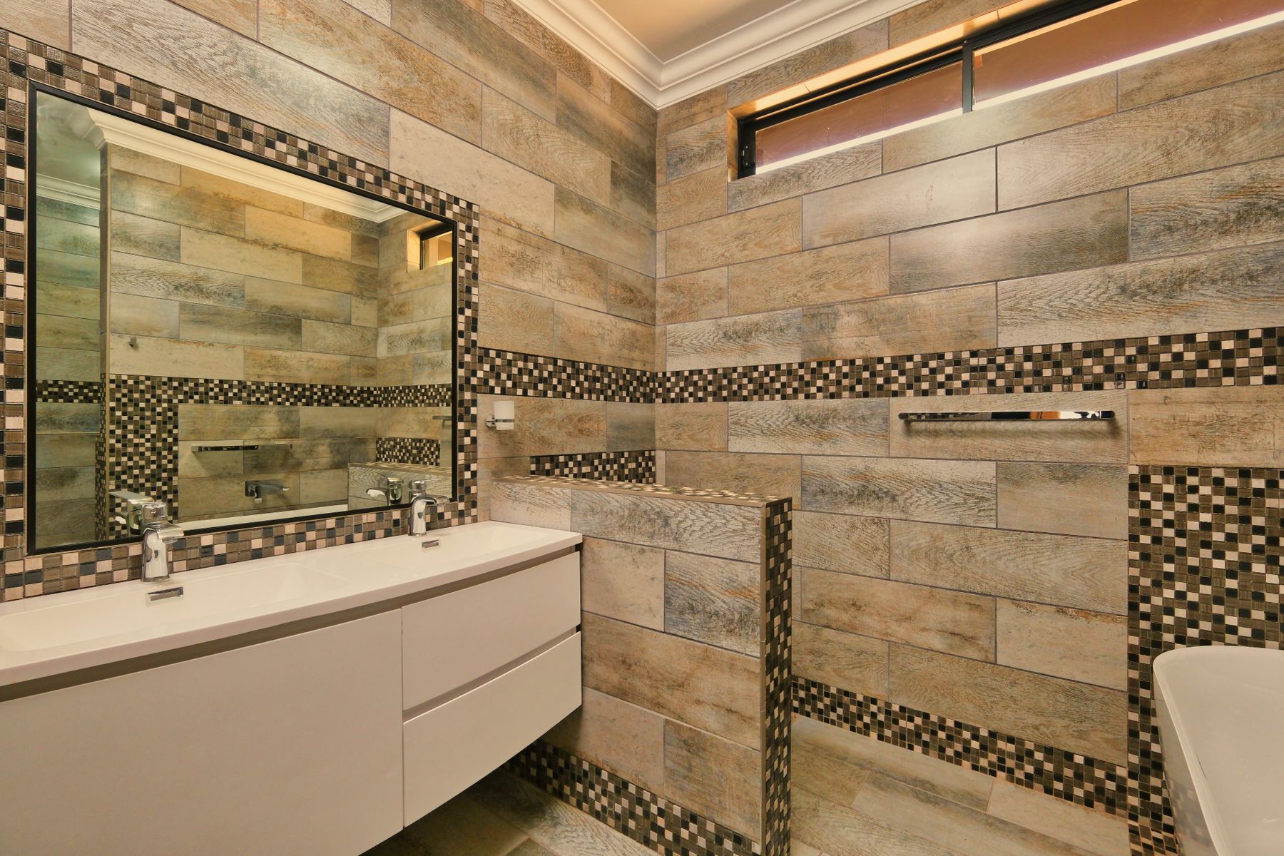 A bathroom clad in horizontal stone tiles with a mosaic band, a white vanity and a bath at the right.