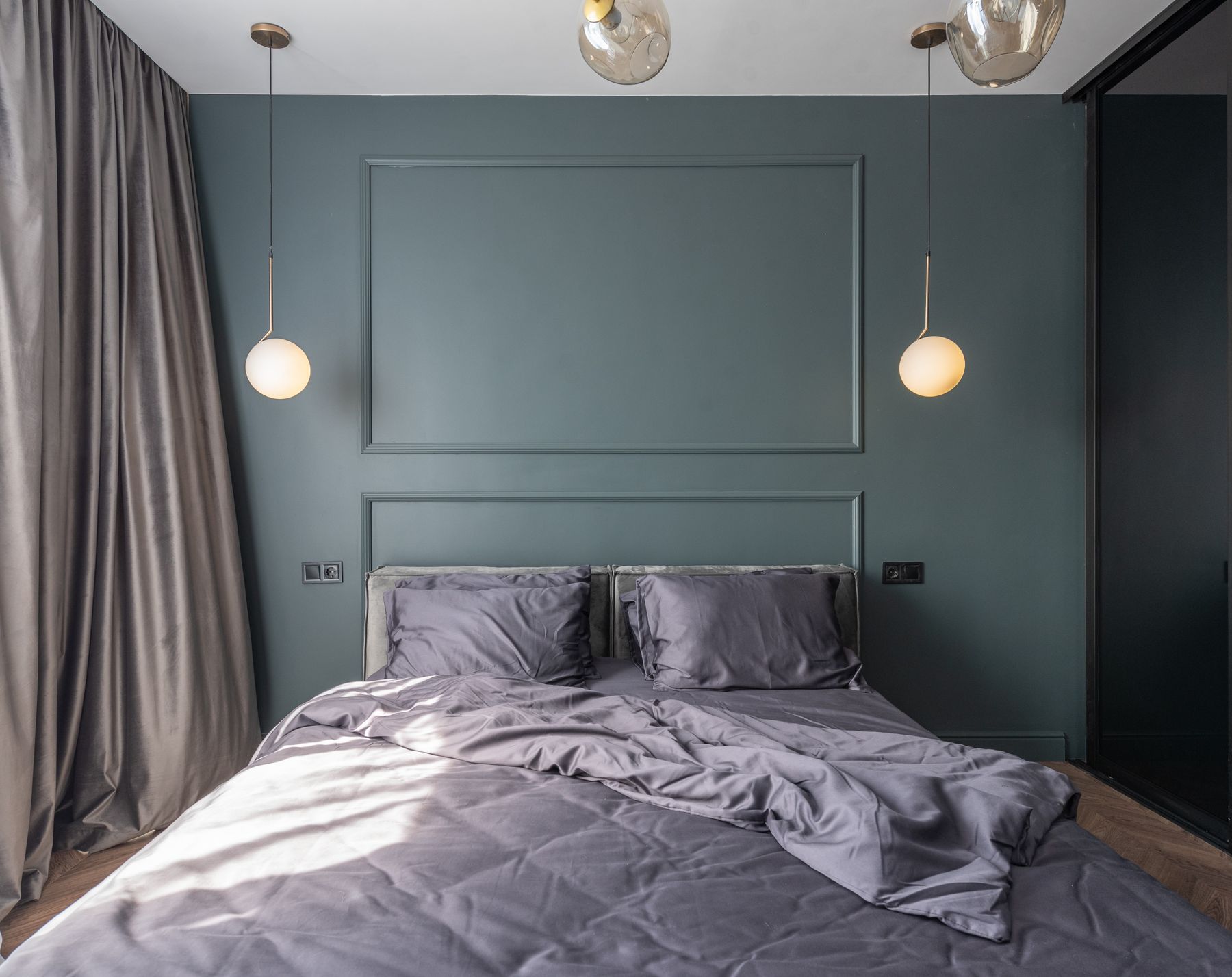 A panelled bedroom painted a deep blue-grey, with two globe pendant lights beside a made bed.