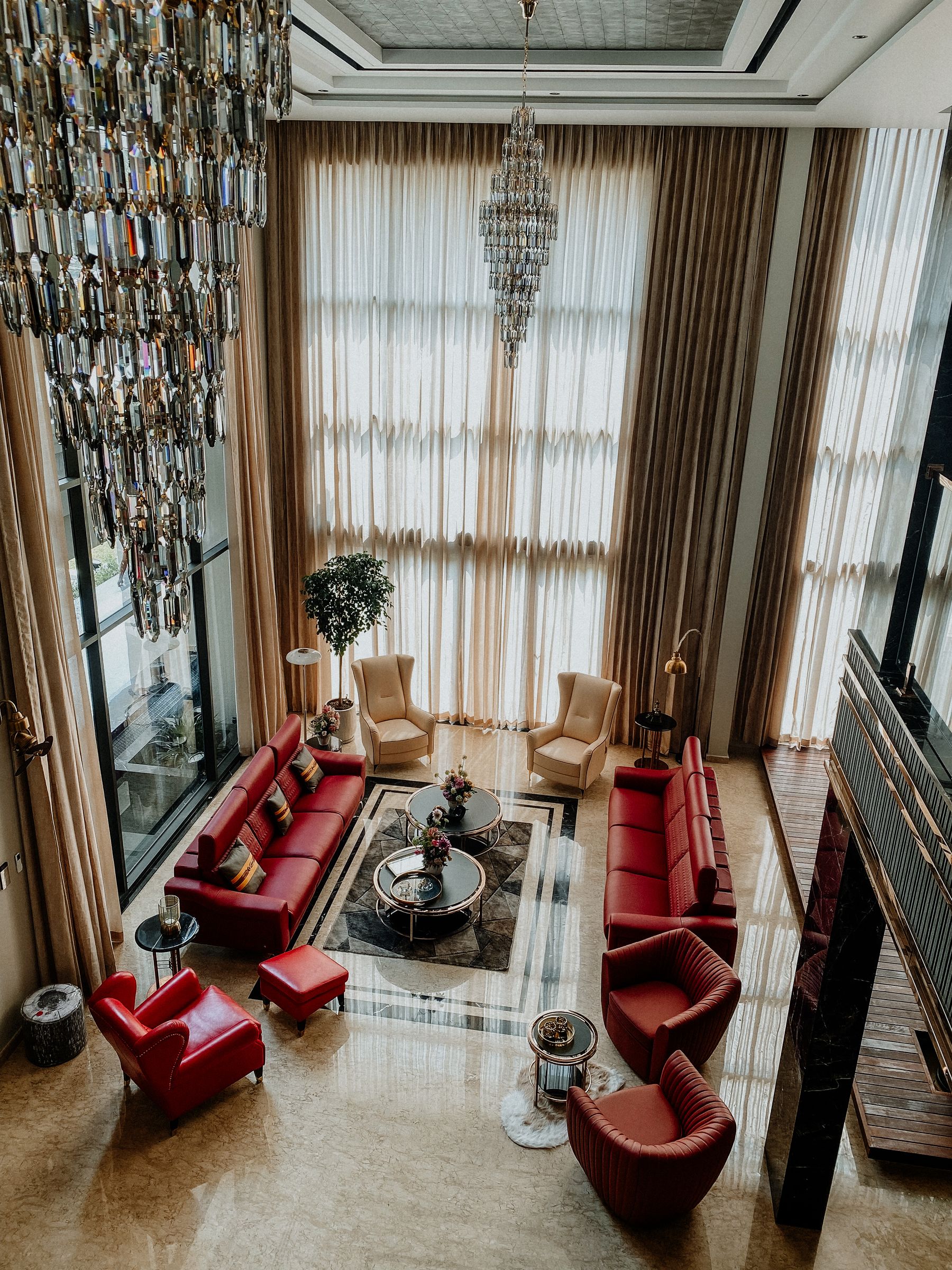 A double-height living room with tall curtains, a chandelier, low seating and a full-height window at the far end.